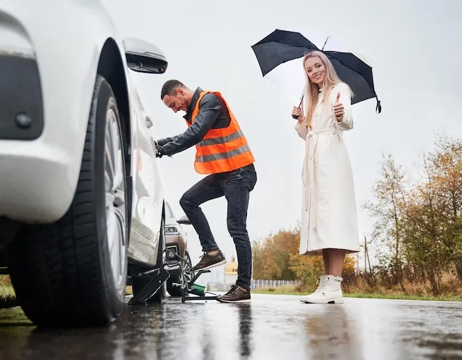 A mechanic in a hi-vis vest changing a wheel on a wet road, beside the woman driver
