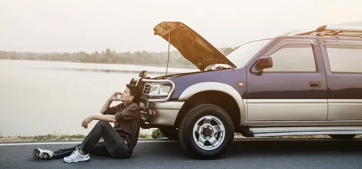 A driver sitting on the tarmac beside a car with its bonnet up, talking on the phone, on a road by a lake